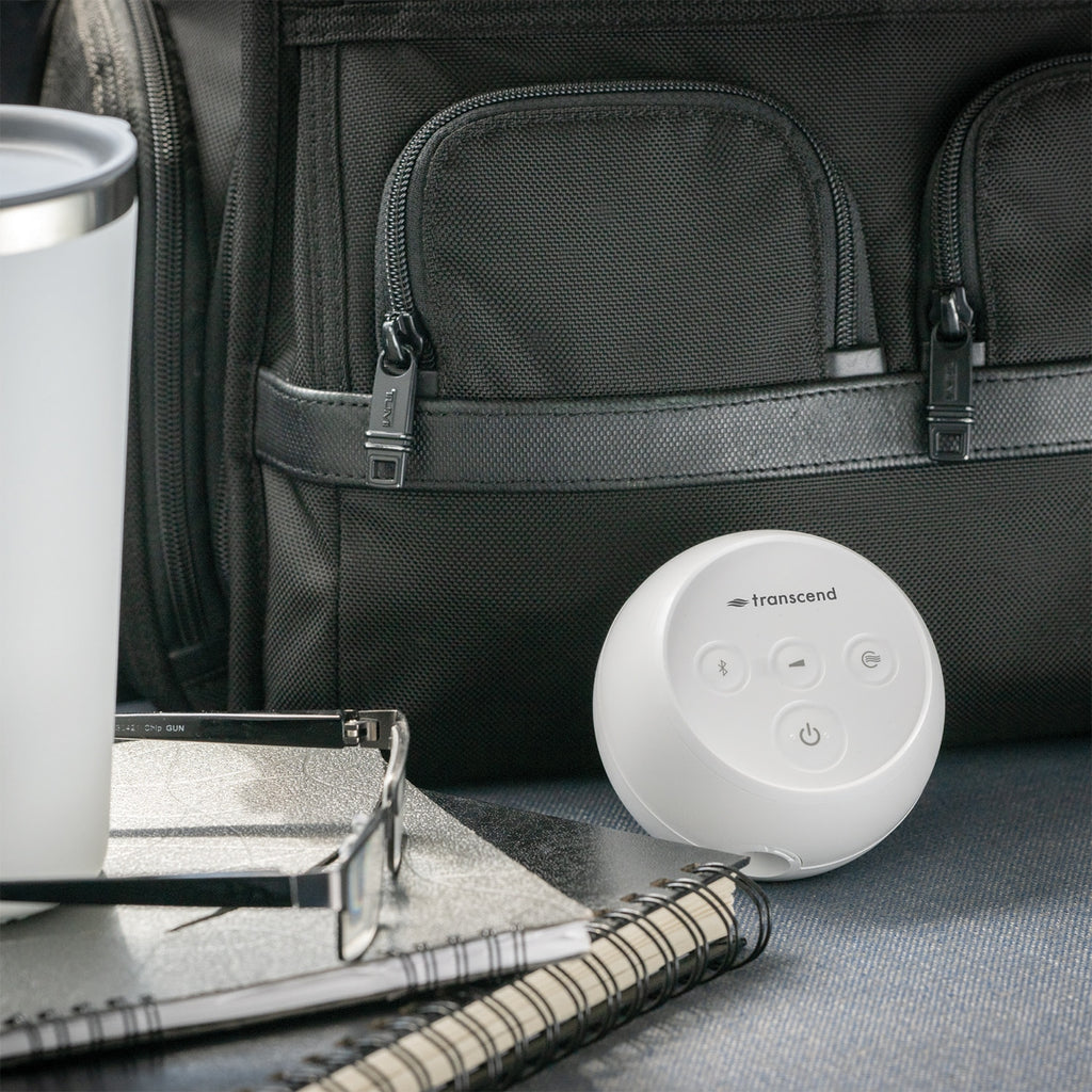 A white Transcend portable Bluetooth speaker placed on a dark textured surface in front of a black backpack, with a white travel tumbler, notebook, and glasses nearby.