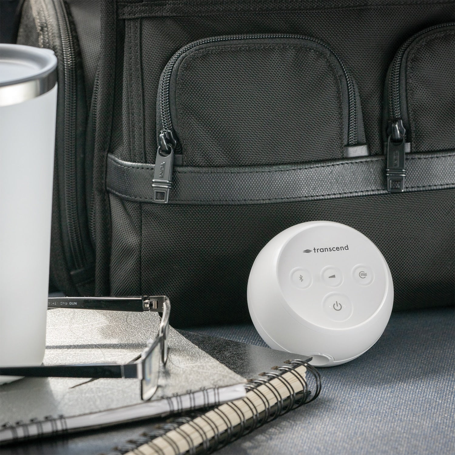 A white Transcend portable Bluetooth speaker placed on a dark textured surface in front of a black backpack, with a white travel tumbler, notebook, and glasses nearby.