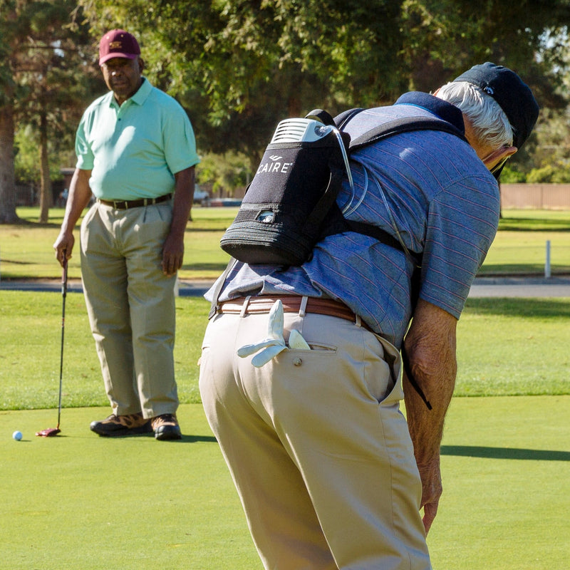 Two men on a golf course, one bending over with a golf club while wearing a Freestyle Concentrator, the other standing and observing.
