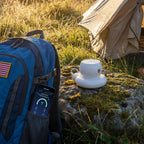 A white portable device sits on a mossy rock beside a blue backpack with a smartphone in the pocket, outdoors near a tent in a grassy field.