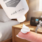 A close-up of a white squeeze tube dispensing a dollop of white cream onto a person's fingertip in a softly blurred bedroom setting.