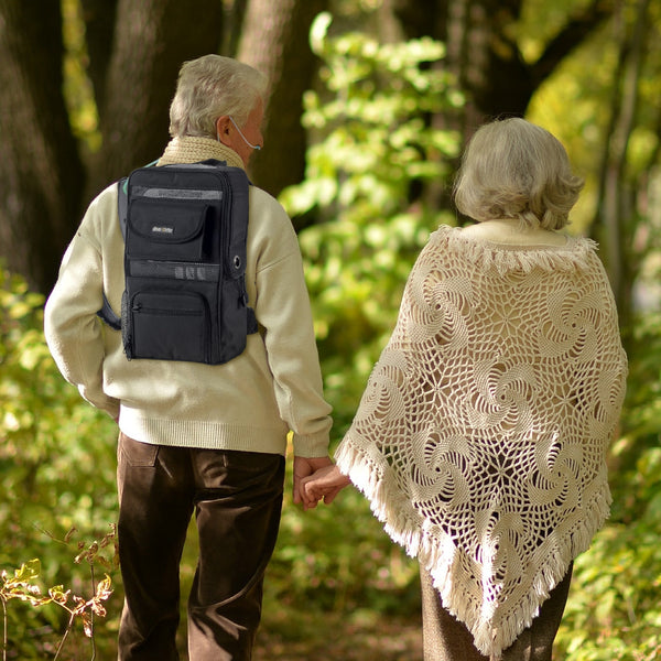 Two elderly people walking hand in hand through a forest, one with a black backpack containing an oxygen concentrator 
