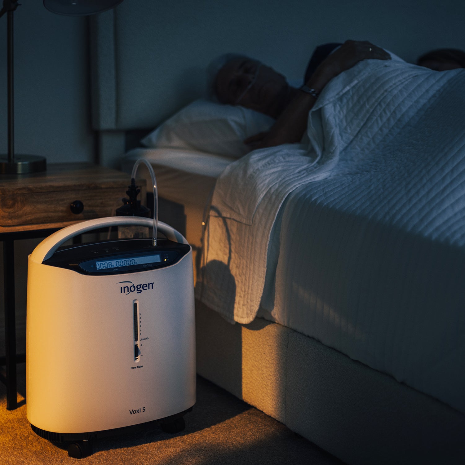 Inogen oxygen concentrator in a bedroom setting with a person lying in bed.