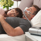 Man using a CPAP machine with a woman lying next to him on a bed.