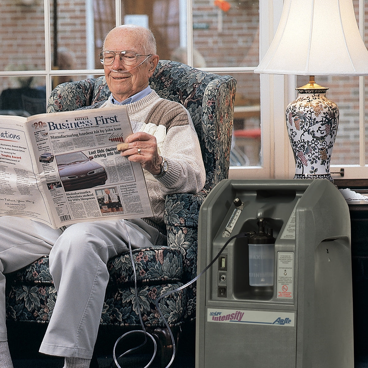 Man reading a newspaper next to an oxygen concentrator in a home setting.