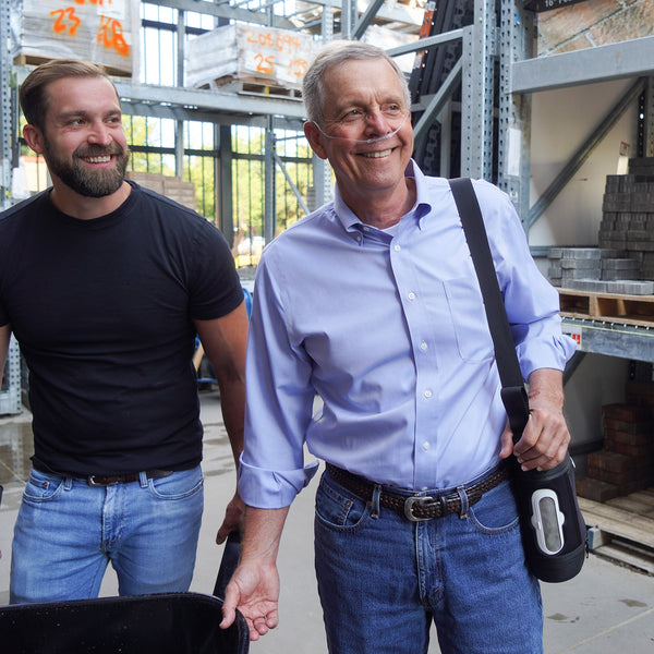 Two men standing in a warehouse setting, one holding a black bag with an Oxlife 2 Portable Oxygen Concentrator 
