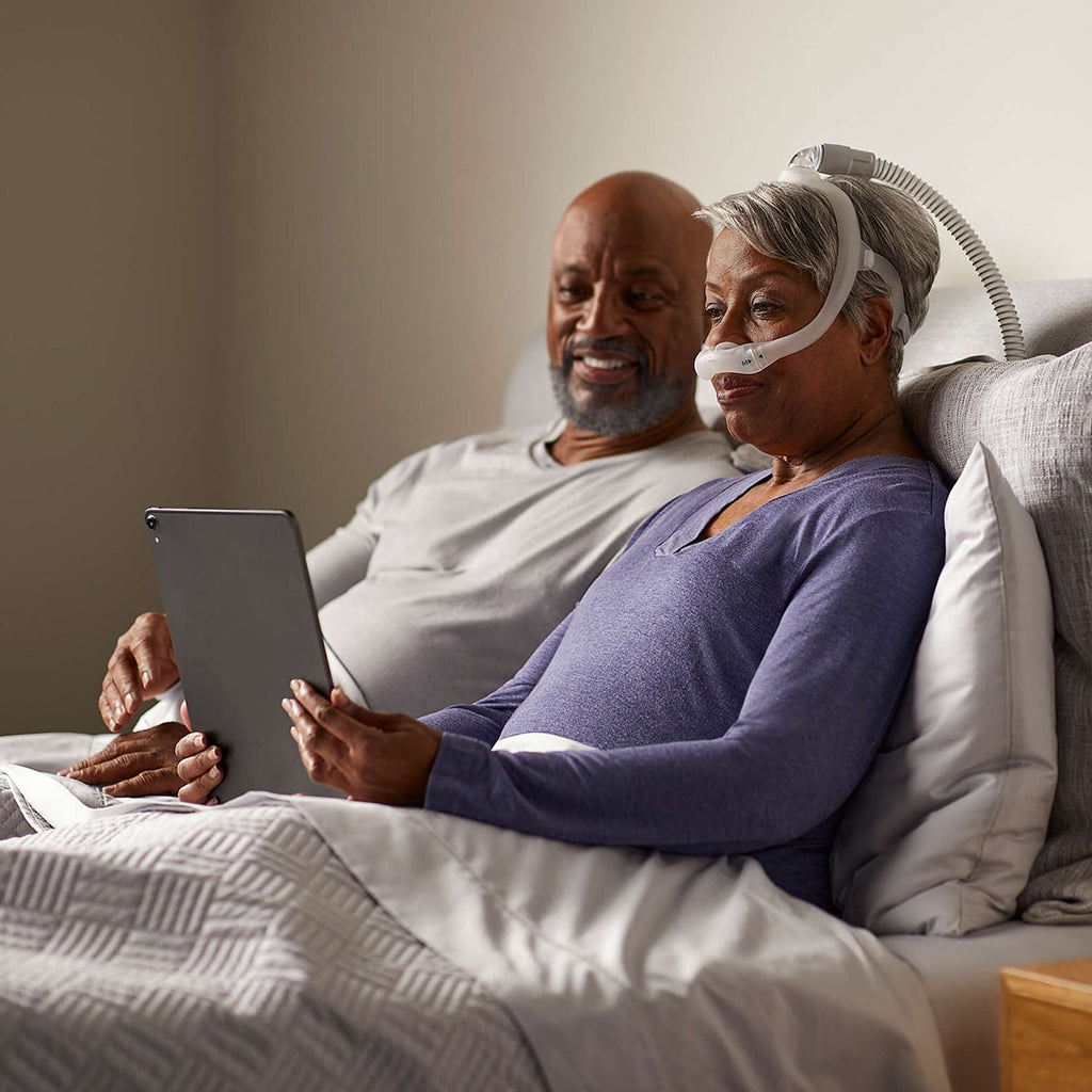 Man and woman in bed, woman using a tablet while wearing a CPAP mask 