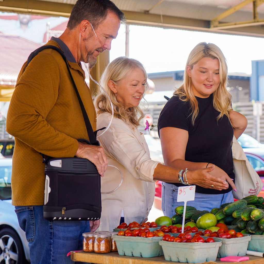 Three people shopping at an outdoor farmers market with produce on display. Man is wearing an Oxlife 2 Portable Concentrator 