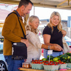 Three people shopping at an outdoor farmers market with produce on display. Man is wearing an Oxlife 2 Portable Concentrator 