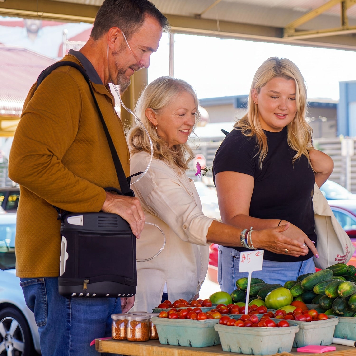 Three people shopping at an outdoor farmers market with produce on display. Man is wearing an Oxlife 2 Portable Concentrator 