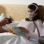 Woman using a CPAP mask and reading a book in bed with a man.